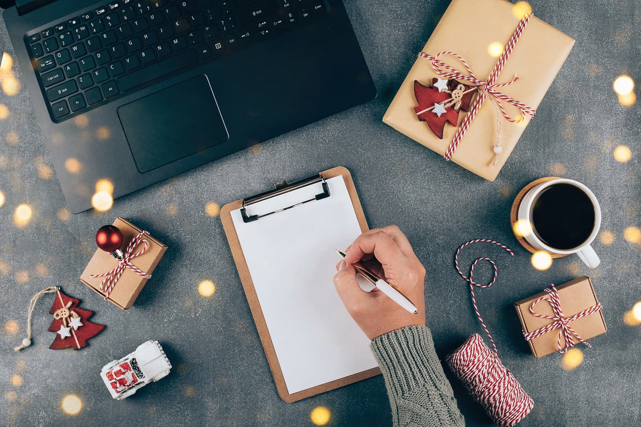 Christmas online shopping flat lay. Girl writing shopping list. Laptop, present box, cup of coffee, holiday decoration. Winter holidays sales
