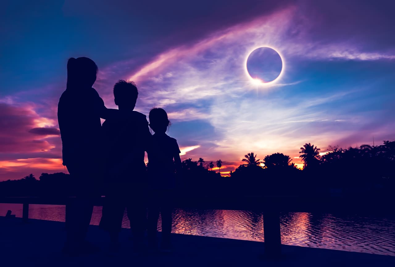 Amazing scientific natural phenomenon. The Moon covering the Sun. Silhouette of mother and children looking at total solar eclipse with diamond ring effect on sky. Happy family spending time together.