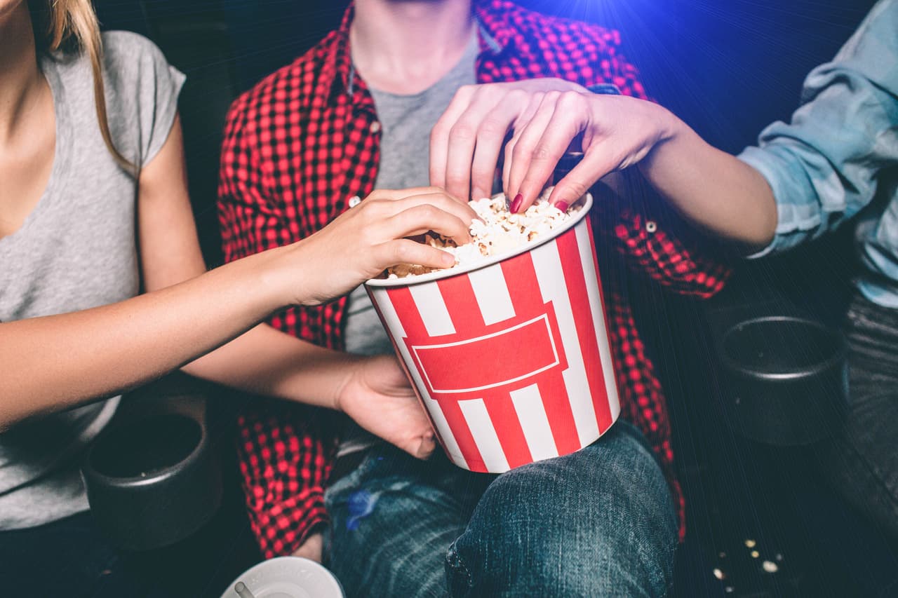 Close up of red with white basket of popcorn that both girl and guy are holding. All of them are taking popcorn out of basket. Сut view